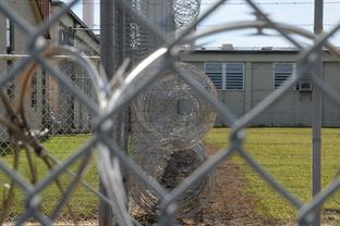 Photograph of a prison seen through a barbed wire fence.