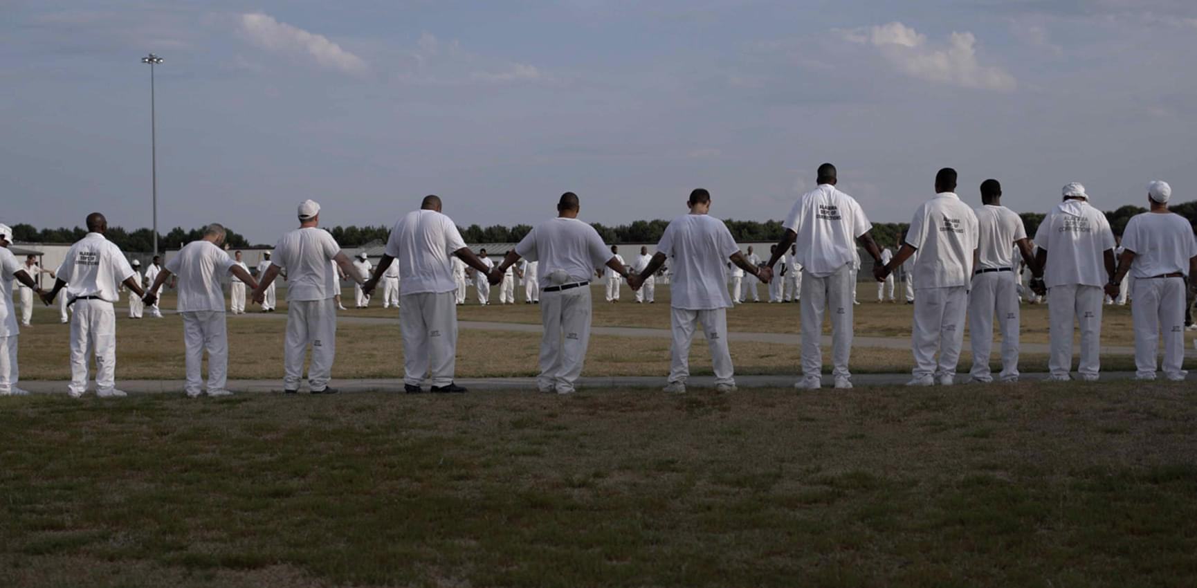 Prisoners dresssed in white uniforms holding hands forming a circle in a prison yard.